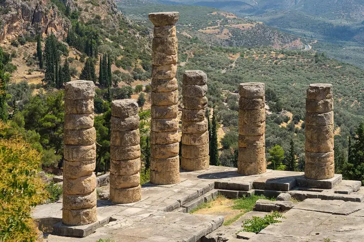 Seis colunas do templo de Apolo em Delfos numa encosta com oliveiras e montanhas ao fundo